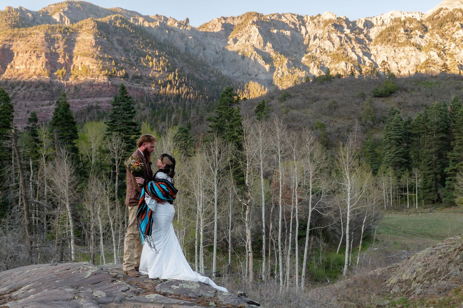 Couple embracing on rocky cliff with mountain forest backdrop during outdoor wedding ceremony.