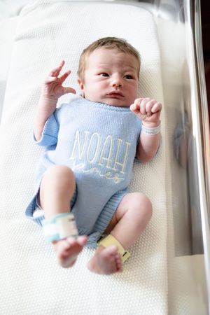 Newborn baby Noah in blue knit onesie lying in hospital bassinet with identification bands on wrists and ankles.