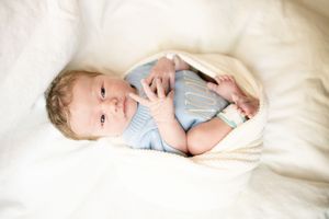 Newborn baby in light blue onesie lying on white blanket, looking up with curious eyes.