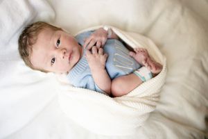 Newborn baby in blue knit onesie wrapped in cream blanket lying on white bedding.