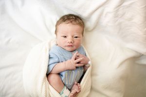 Newborn baby in blue onesie wrapped in cream blanket lying on white bedding, peaceful infant portrait.