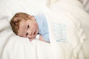 Newborn baby wrapped in a personalized white knit blanket embroidered with the name Noah.