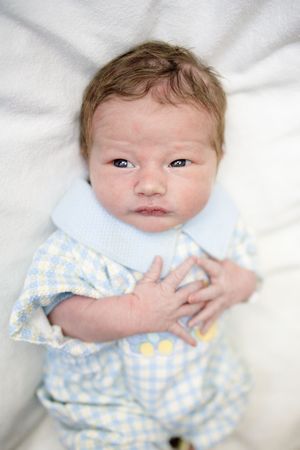 Newborn baby in blue checkered outfit lying on white blanket, looking alert with dark eyes.