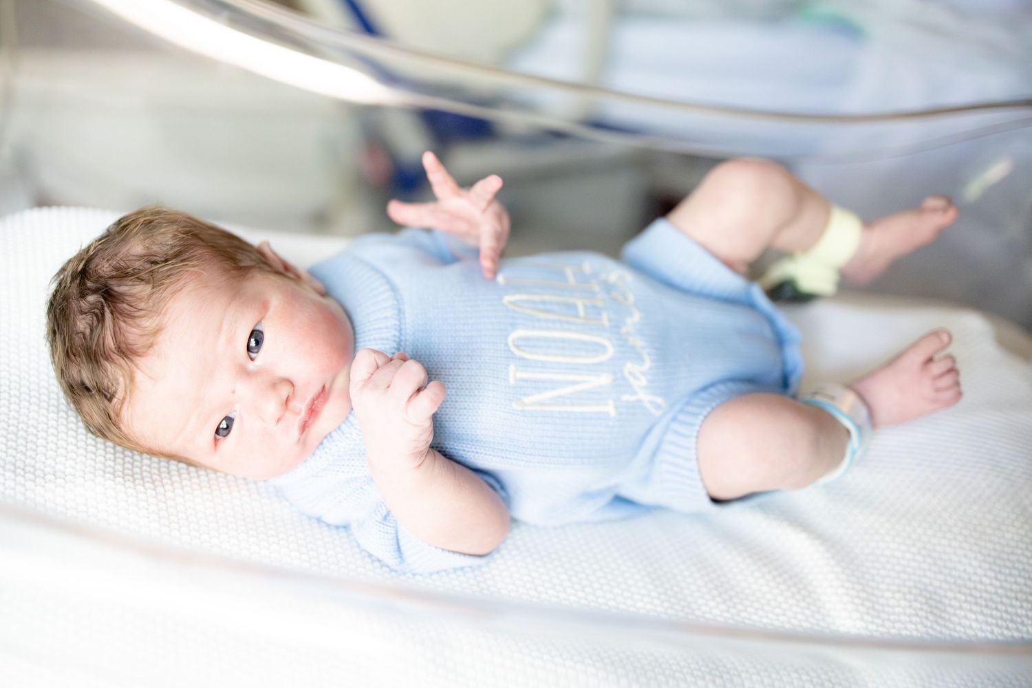 Newborn baby in blue onesie lying in hospital incubator under medical lighting.