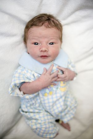 Newborn baby in blue checkered onesie lying on white blanket, looking at camera with alert dark eyes.