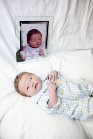 A baby in blue plaid onesie lies on white fabric beside a framed newborn photo, showing growth over time.