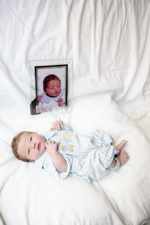 Newborn baby in blue outfit lying on white bedding next to a framed photo of another infant.