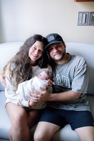 Happy couple sitting on a couch smiling while holding their newborn baby wrapped in a white blanket.
