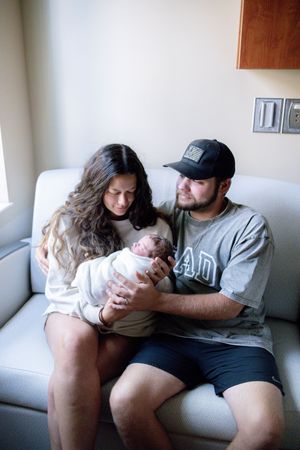 New parents sitting on a couch together, lovingly gazing at their newborn baby wrapped in a white blanket.