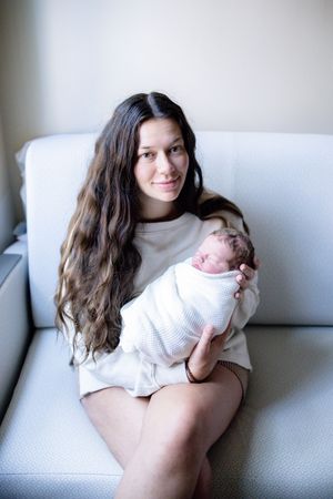 Young mother with long dark hair sits on a gray sofa, gently cradling her swaddled newborn baby.