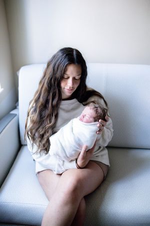 A young mother with long brown hair sits on a white sofa, gently cradling her swaddled newborn baby.
