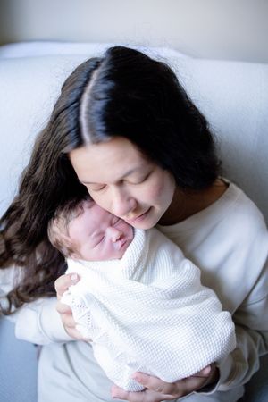 A mother with dark curly hair gently kisses her swaddled newborn baby wrapped in a white blanket.