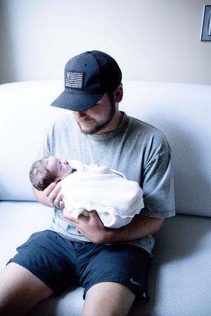 Young man in baseball cap tenderly holds swaddled newborn baby while sitting on a white couch.