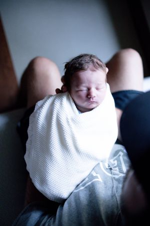 Newborn baby swaddled in white blanket sleeping peacefully on parent's lap in soft natural light.