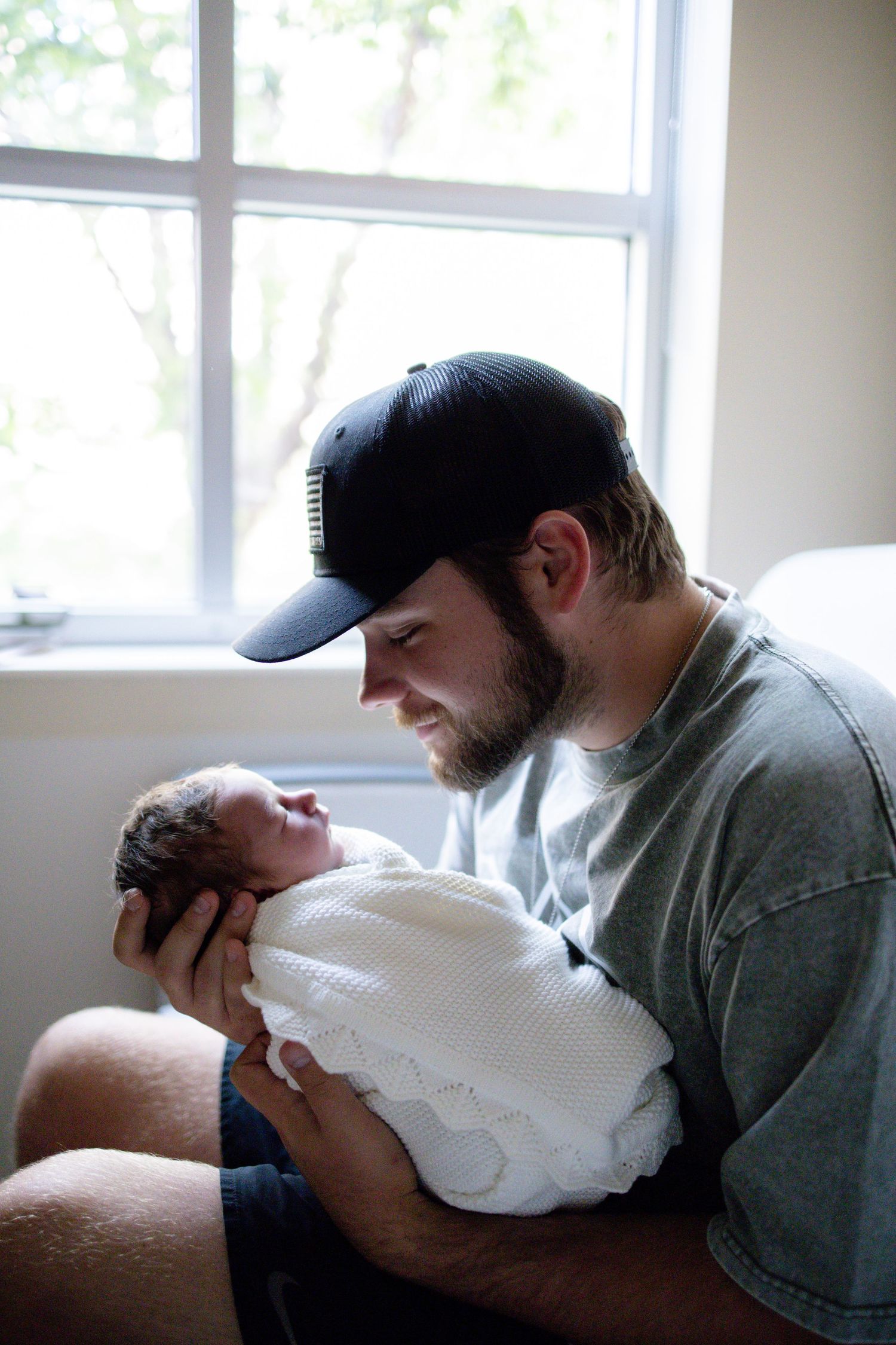 A bearded man in a black cap tenderly holds a sleeping newborn baby wrapped in white near a bright window.