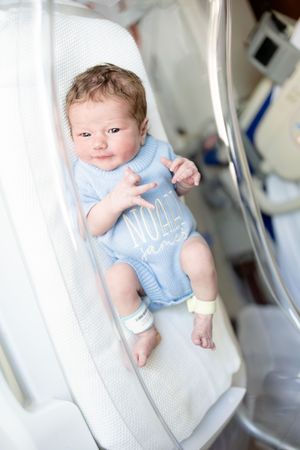Newborn baby in blue onesie lying in hospital bassinet with ID bracelet on wrist.