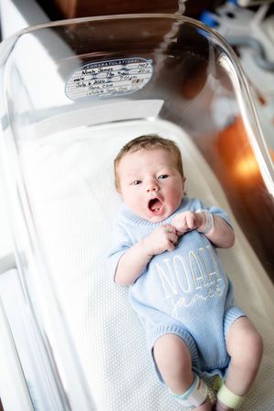 Newborn baby named Noah James yawning in hospital bassinet, wearing a blue knit romper with name embroidered.