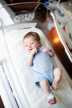 Newborn baby boy named Noah James lying in hospital bassinet wearing a blue knit romper with his name embroidered on it.