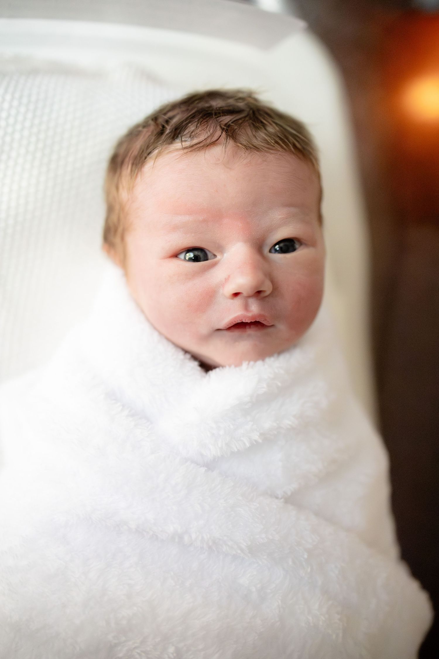 Newborn baby swaddled in a soft white blanket, with dark eyes open, lying in a white bassinet.