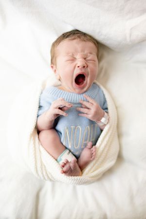 Newborn baby named Noah yawning in a blue knit onesie, wrapped in a cream blanket on a white surface.