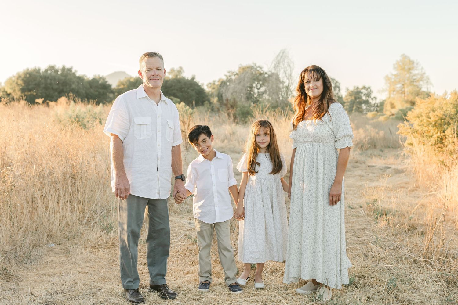 Family of four posing in golden field, parents holding hands with two children in white outfits during sunset.