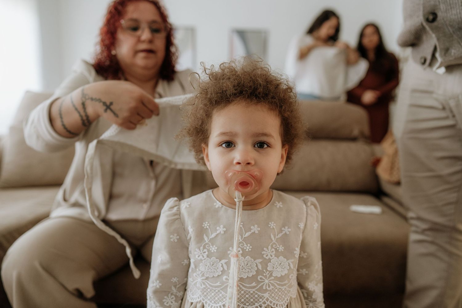 A toddler girl in a white lace dress blows bubbles while a woman combs her curly hair indoors.