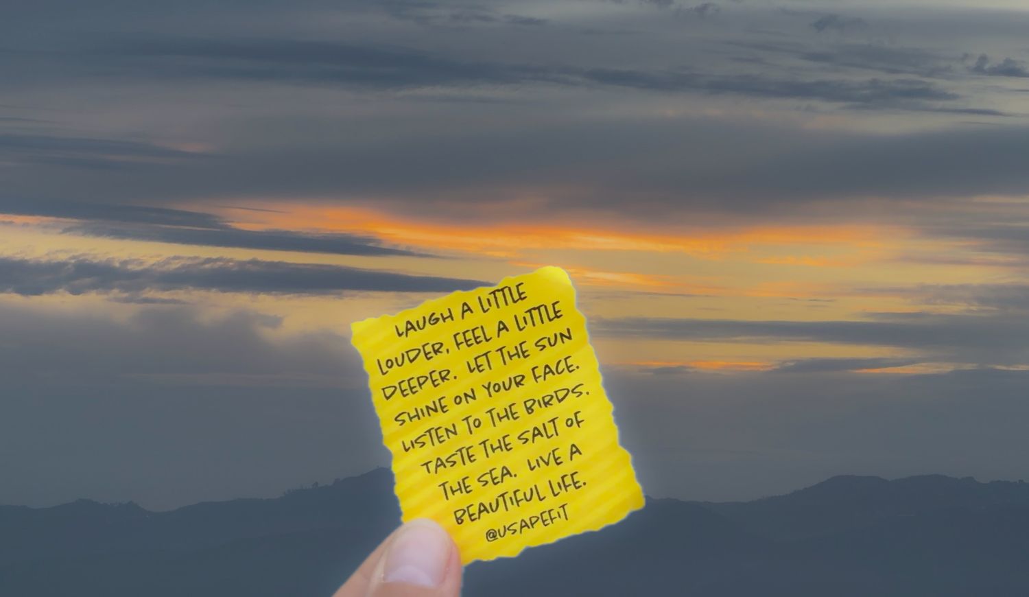 Hand holding a yellow sticky note with inspirational quote against a dramatic sunset sky with orange and gray clouds.