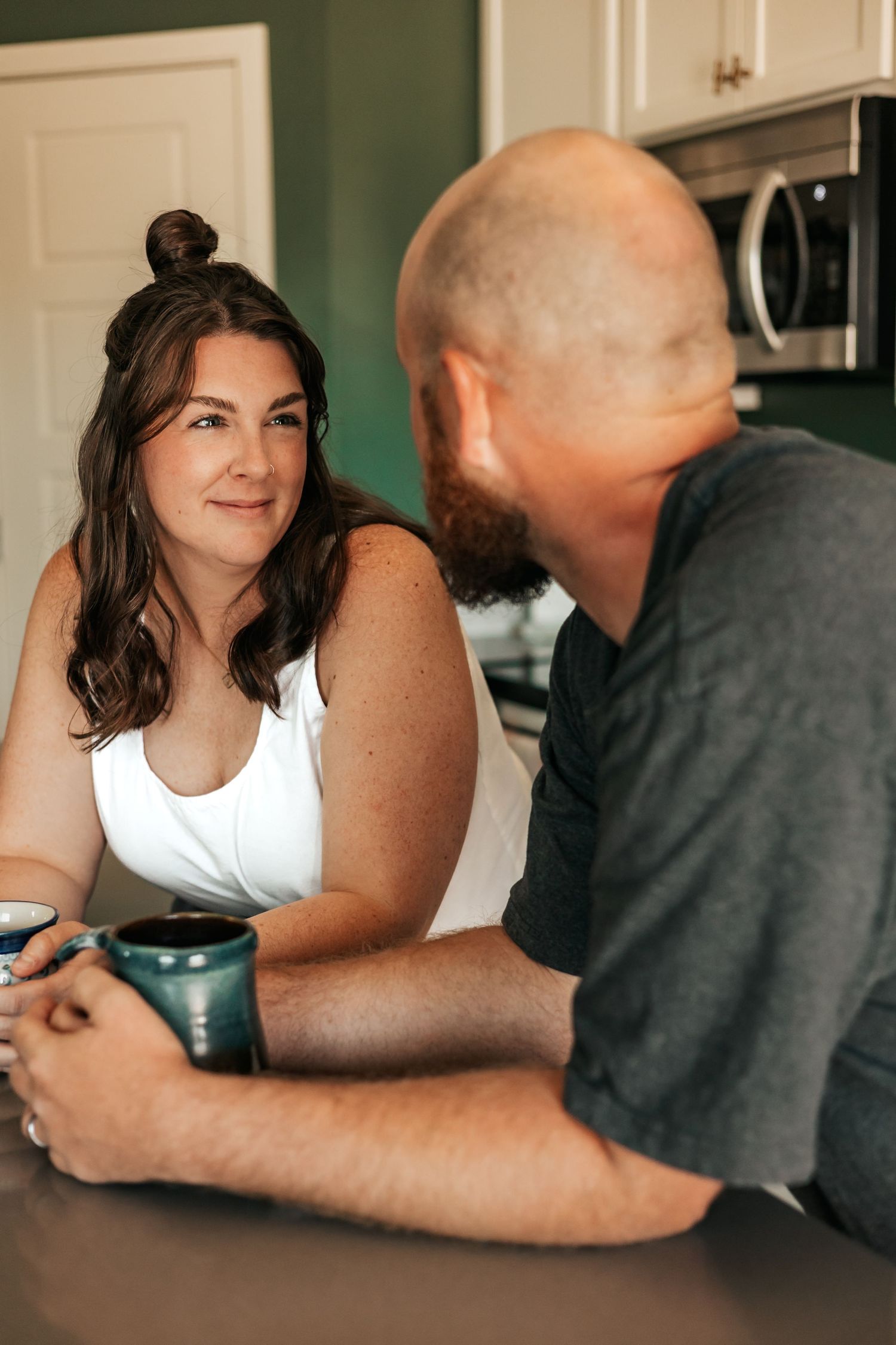 A woman with long brown hair holds a teal mug while chatting with a bald bearded man in a kitchen | Anniversary Photos in Johnston, Colorado