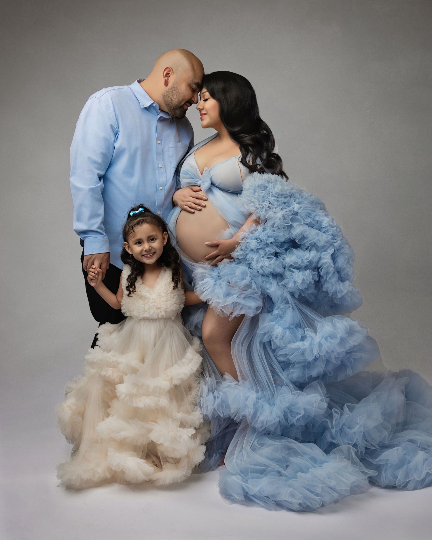 Family maternity photo with pregnant woman in blue tulle gown, man in blue shirt, and young girl in cream dress.