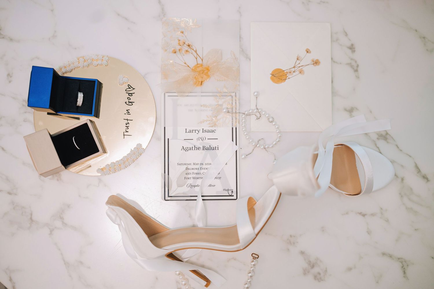 Wedding flat lay featuring invitation suite, white heels, pearl necklace, and ring boxes on marble surface.