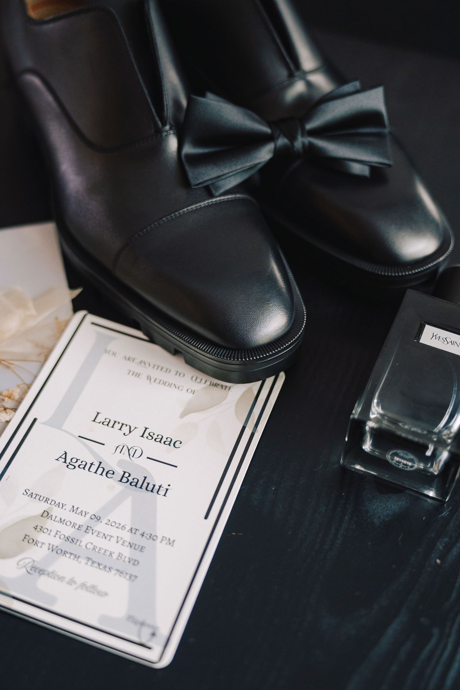Black leather bow-tie dress shoes beside a wedding invitation for Larry Isaac and Agathe Baluti with cologne on dark surface.