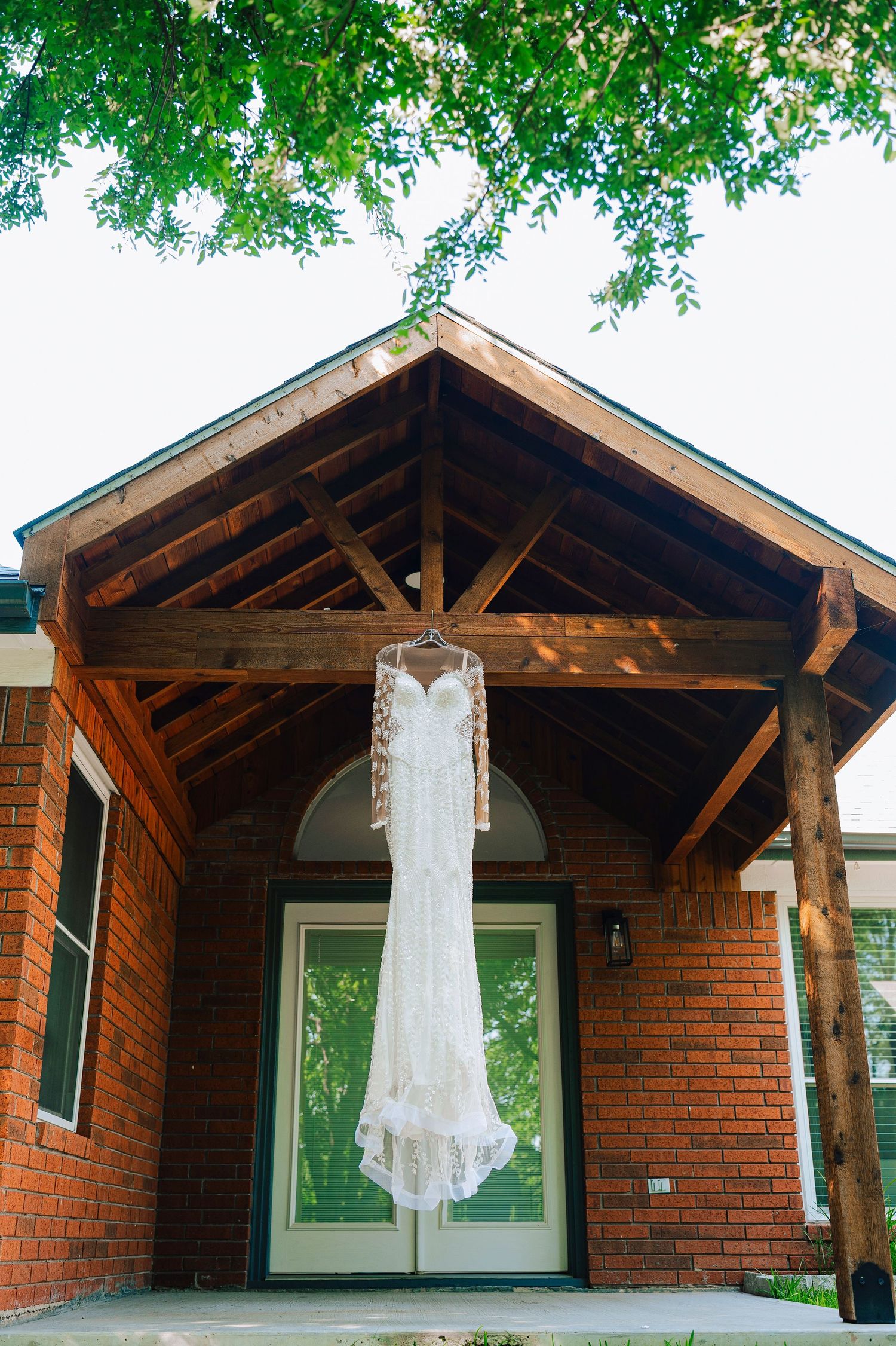 Elegant lace wedding dress with long sleeves hanging on a hanger under a rustic wooden porch of a brick building.