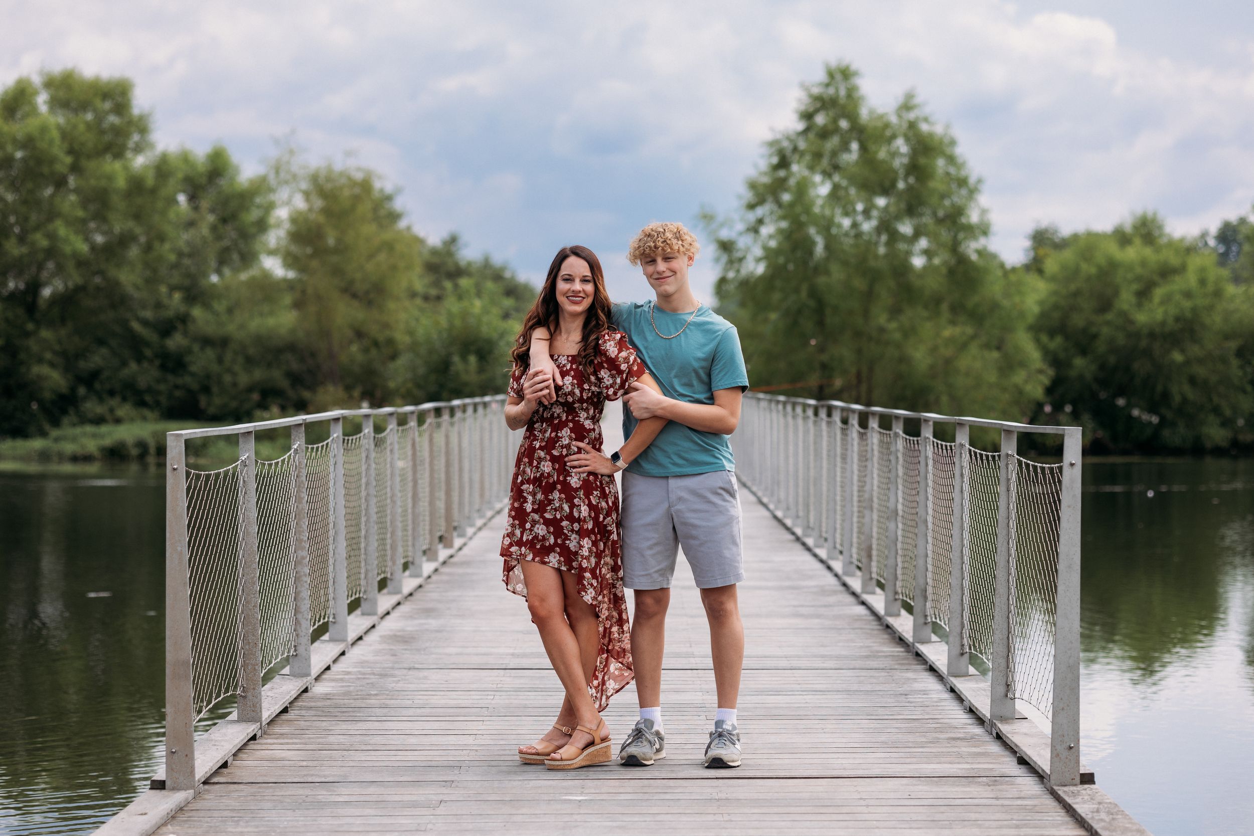 A couple poses on a wooden bridge over a calm lake surrounded by lush green trees on a cloudy day.