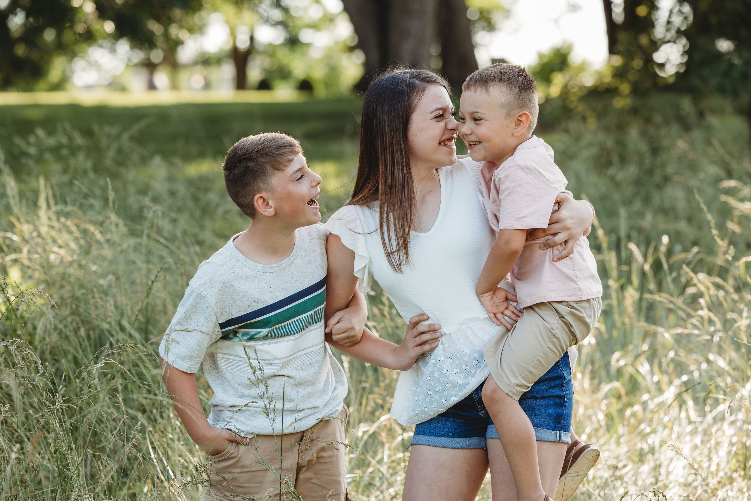 A mother smiles and holds her toddler while her older son looks on in a sunny green field.