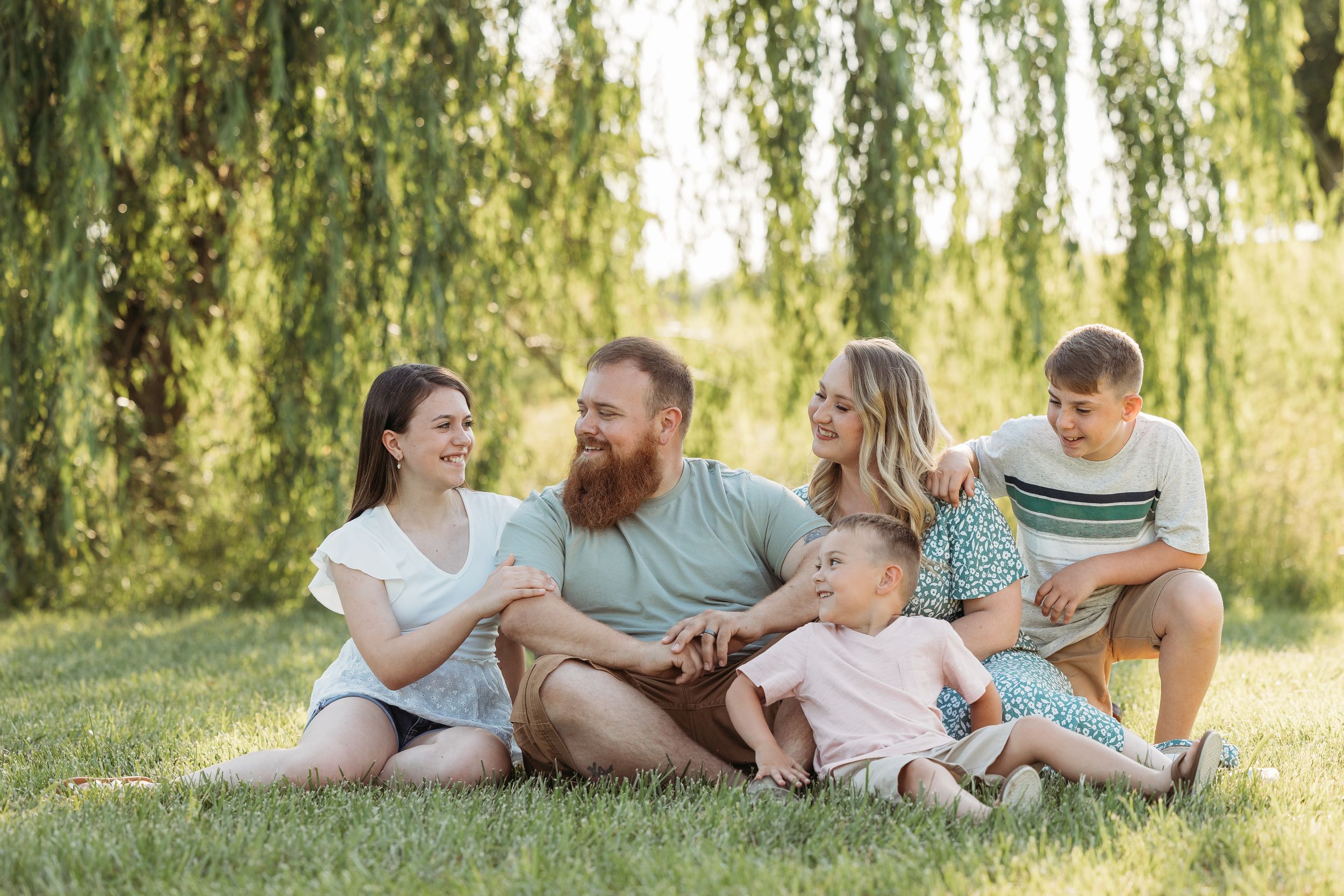 Happy family of five sitting on green grass under willow tree, smiling and enjoying quality time outdoors.