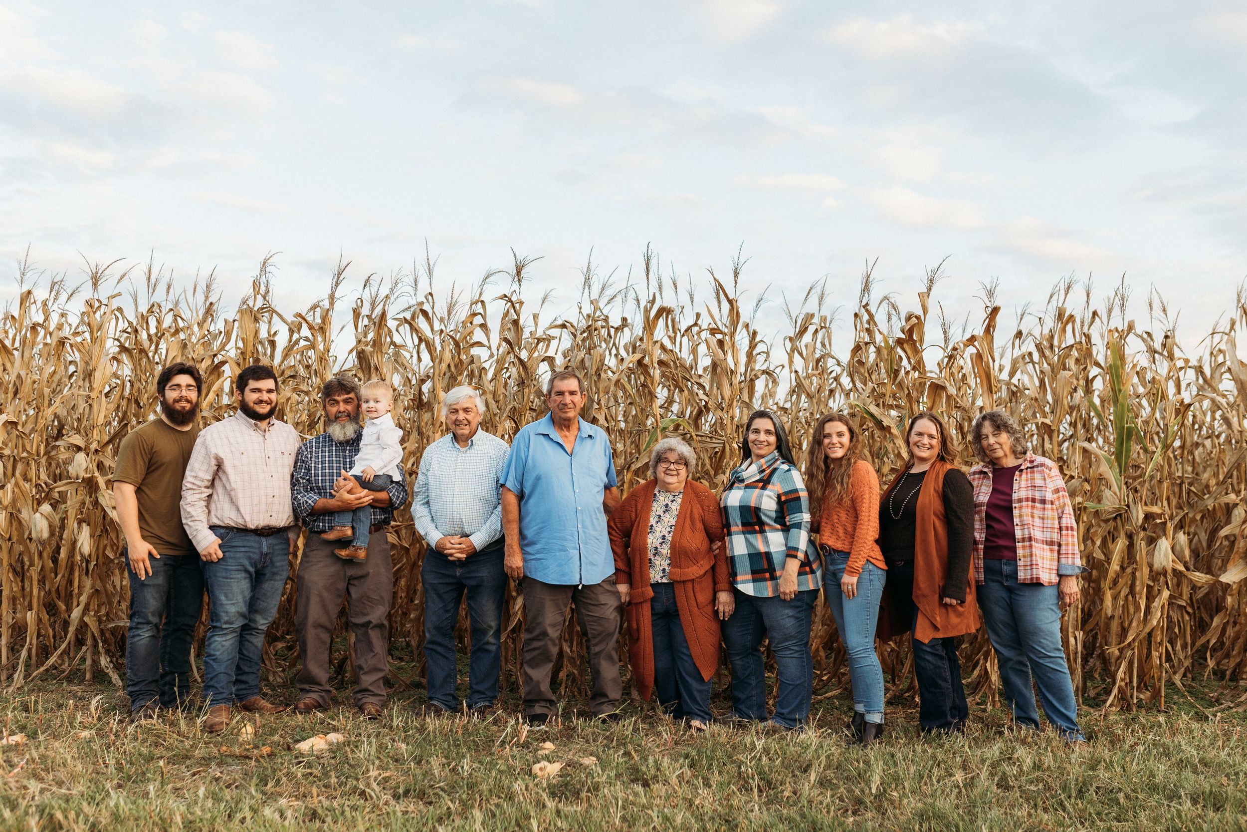 Large family group smiling together in front of tall golden corn stalks in an autumn farm field.