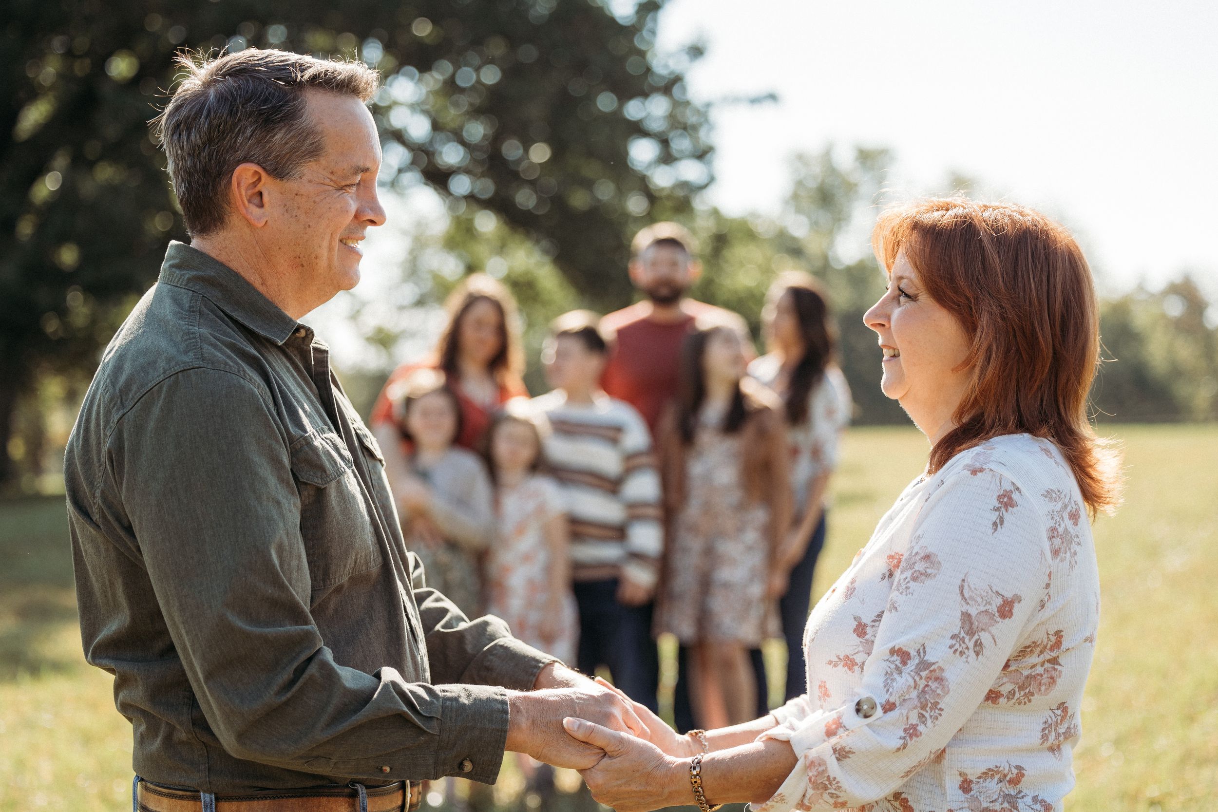 A couple holds hands smiling at each other outdoors, with a group of family members standing together in the background.