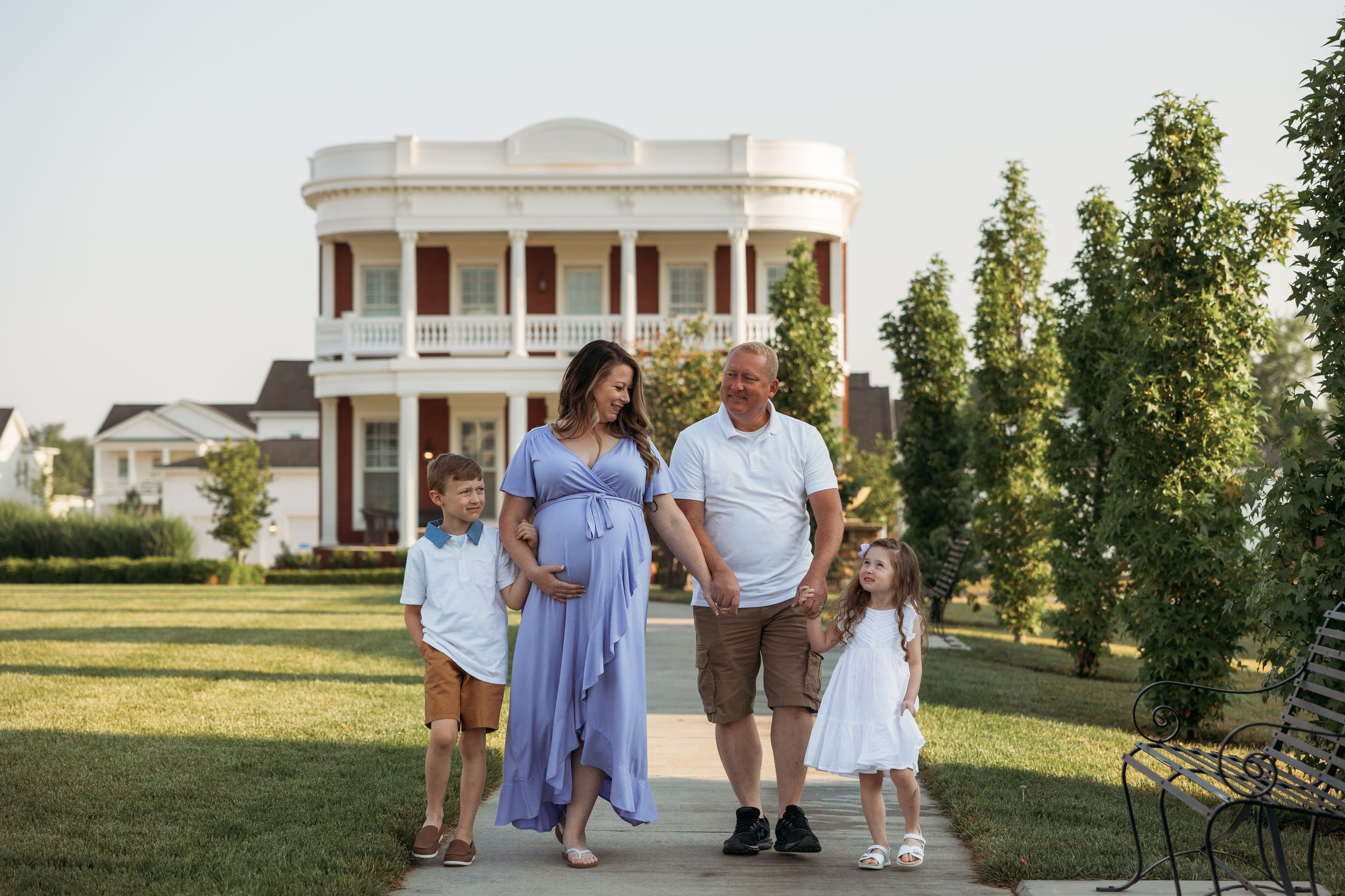 Pregnant woman in blue dress poses with family including two children in front of elegant white Southern mansion.