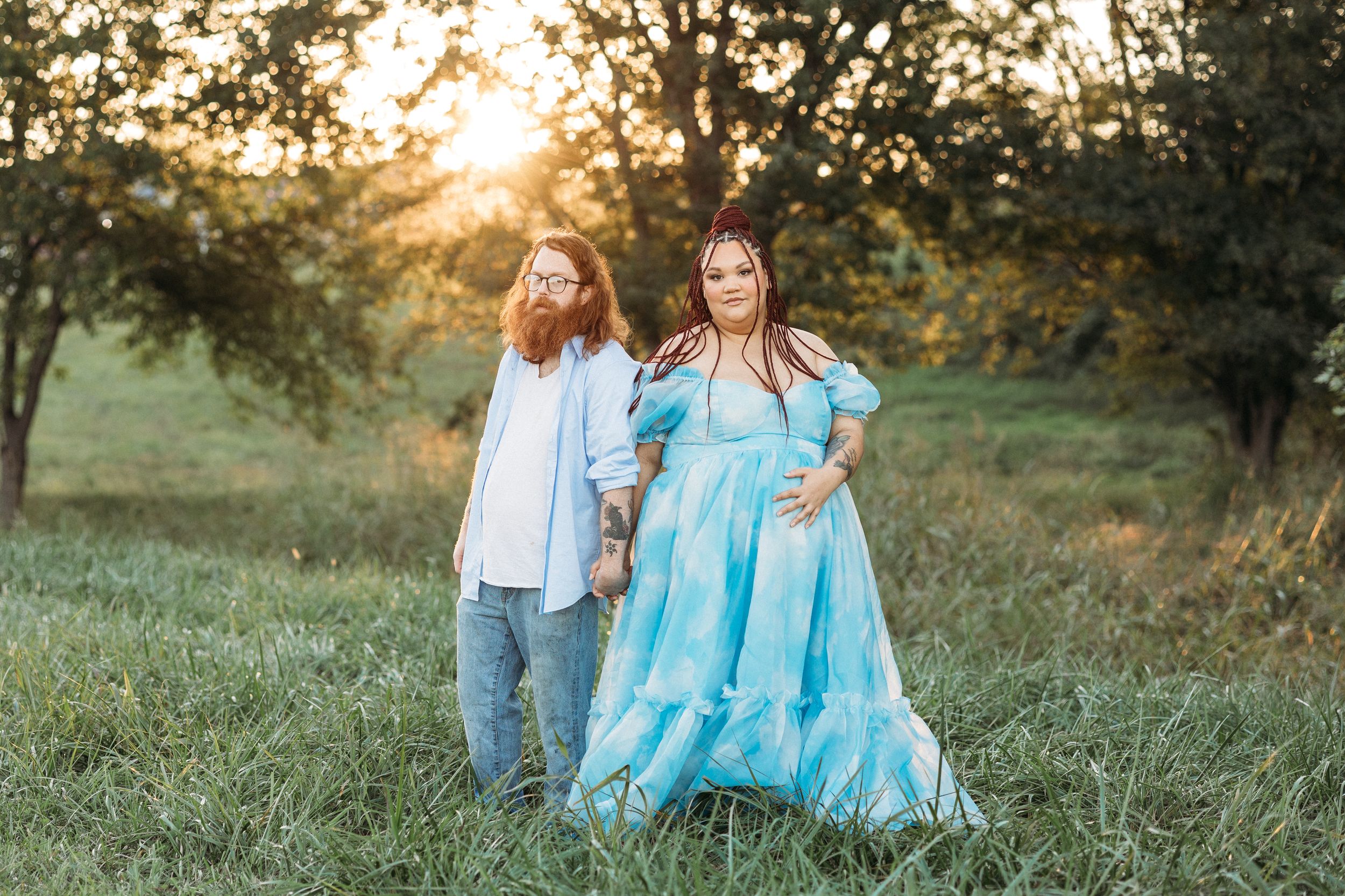 Pregnant couple holding hands in a sunlit field, woman in a flowing teal gown, man with red beard in white shirt.
