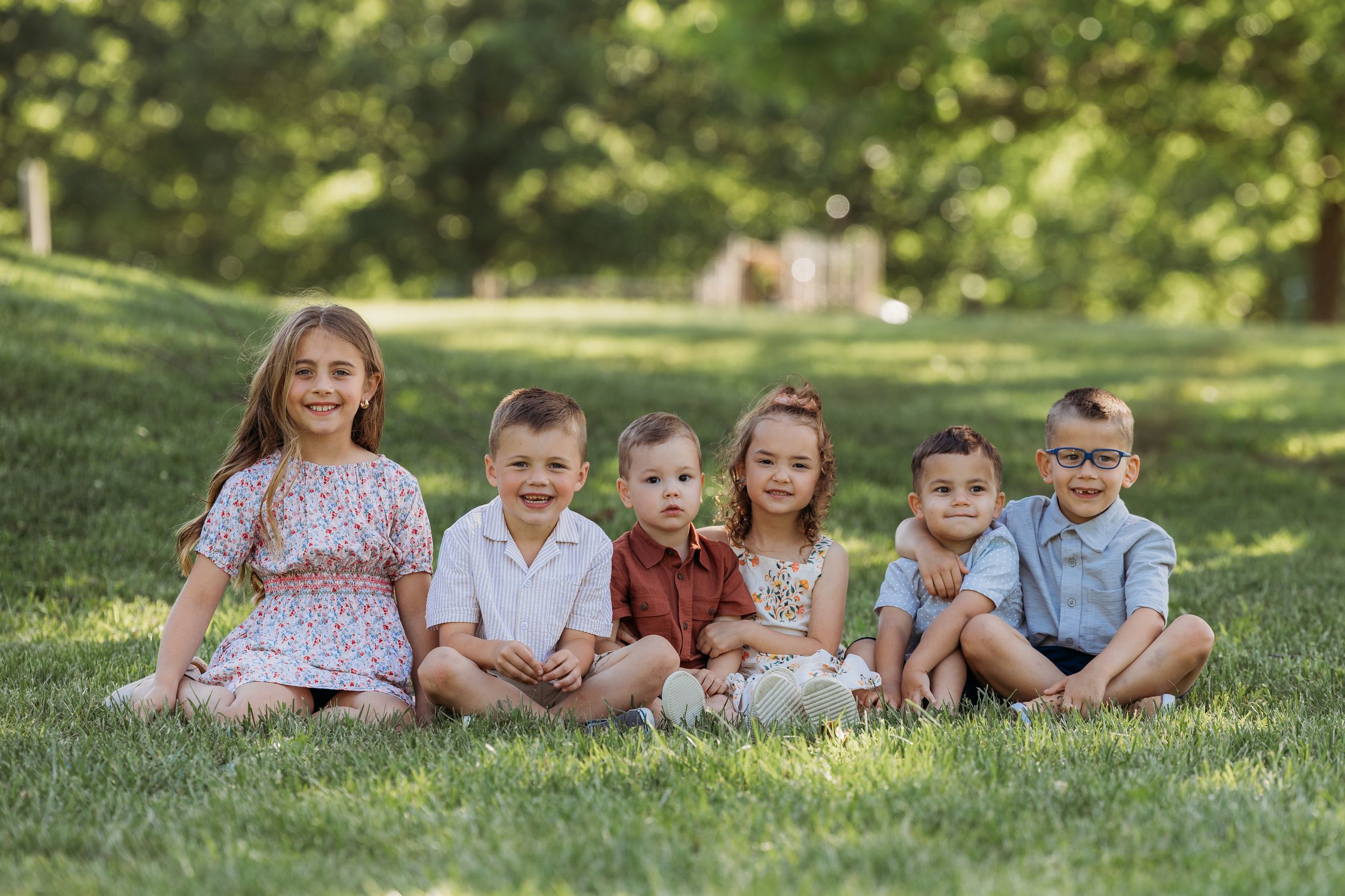 Six young children sitting together on green grass outdoors, smiling in coordinated outfits for a group portrait.