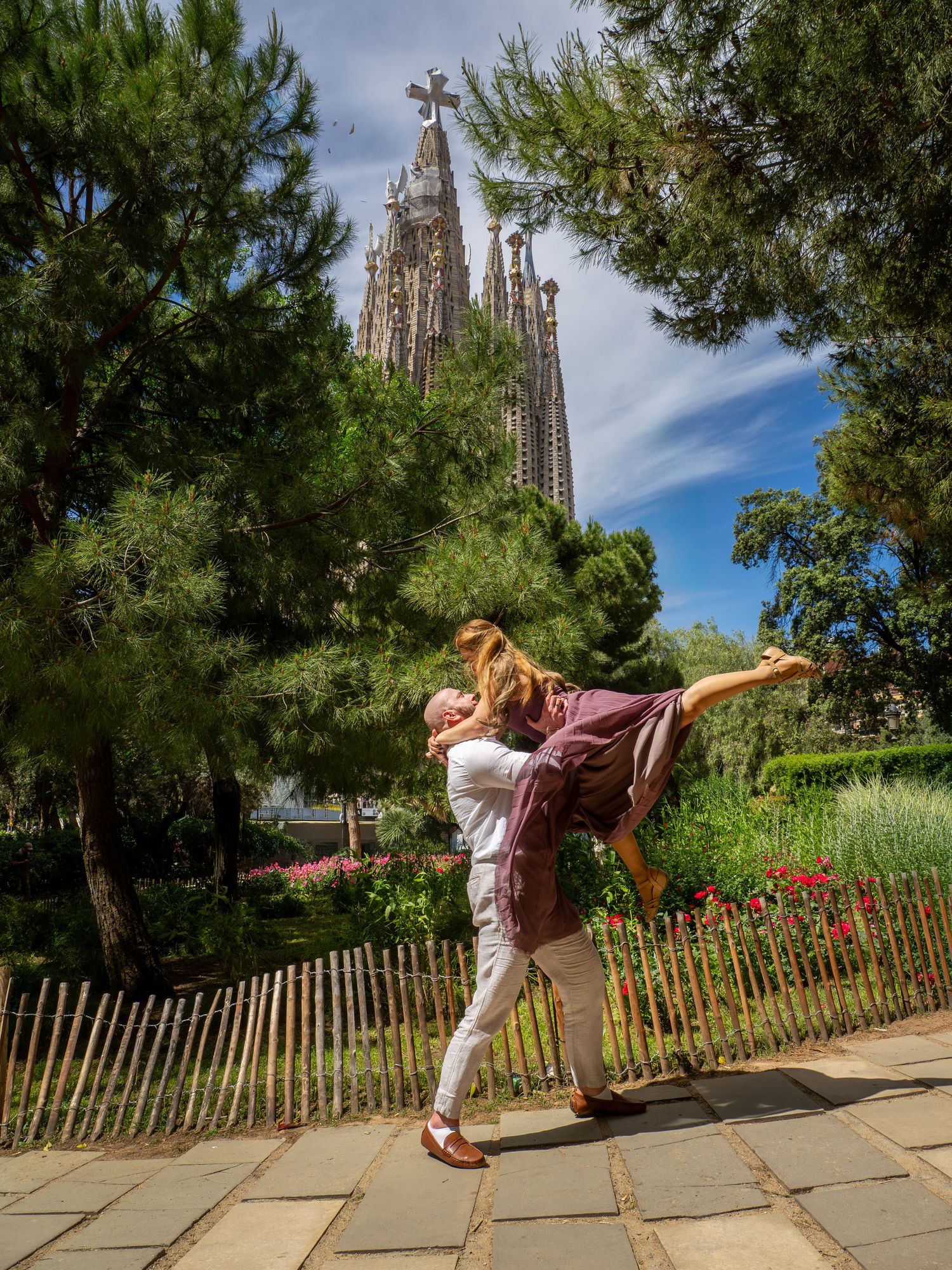 Couple embracing in front of Sagrada Familia basilica in Barcelona, surrounded by lush green trees and gardens.