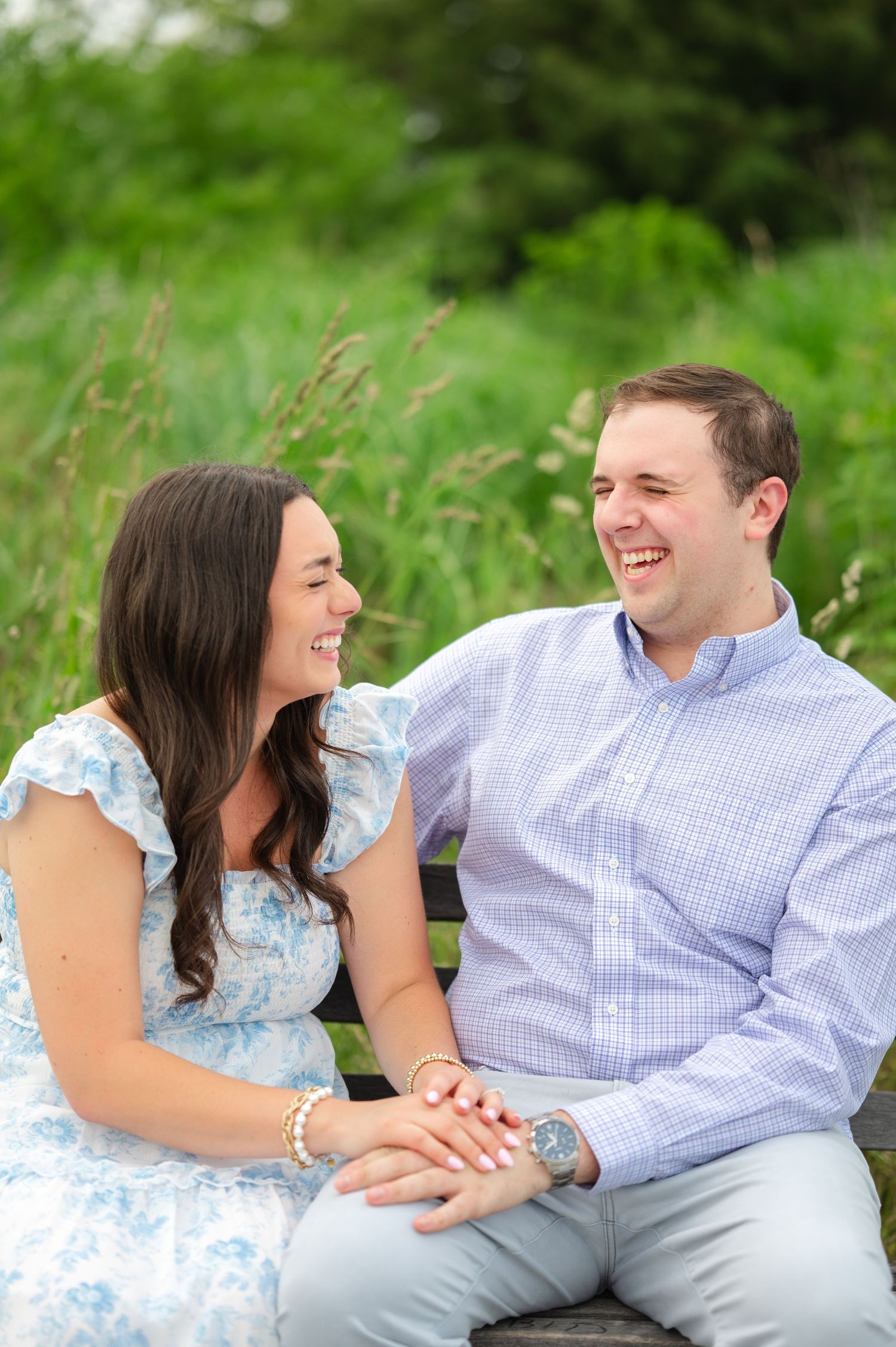 A smiling couple sits on a bench outdoors, laughing together, woman in blue floral dress, man in light blue checkered shirt.