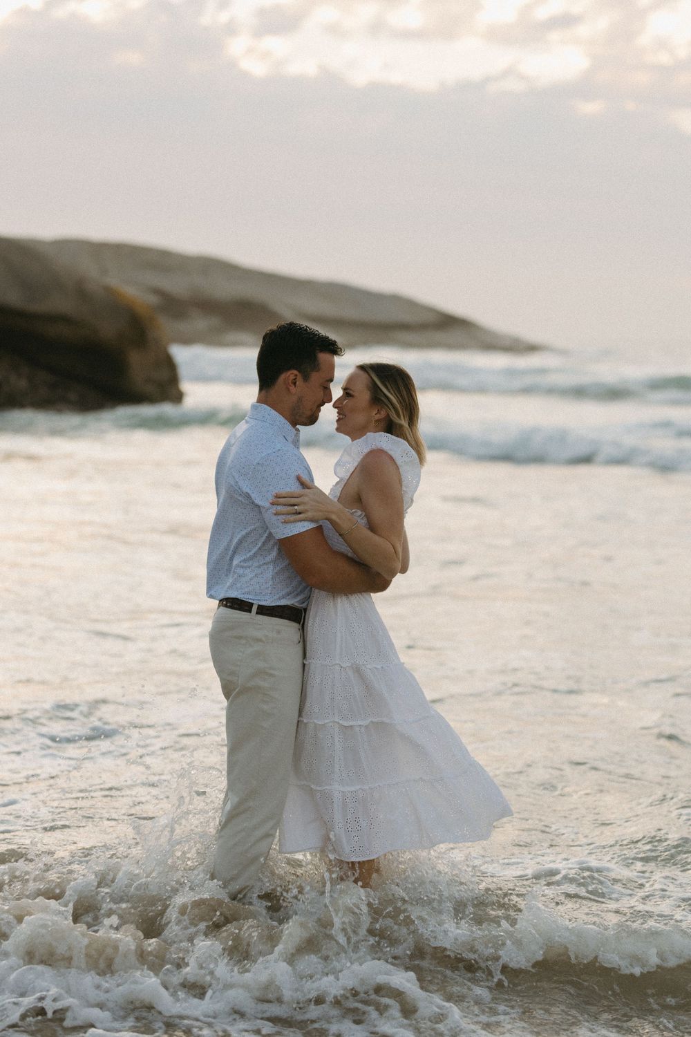 Couple embracing romantically in ocean waves at sunset, woman in white dress, man in light blue shirt on sandy beach.