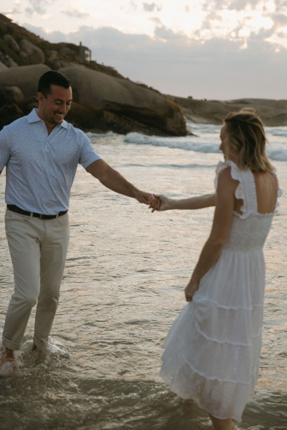 Man and woman holding hands walking through ocean waves on rocky beach at dusk.