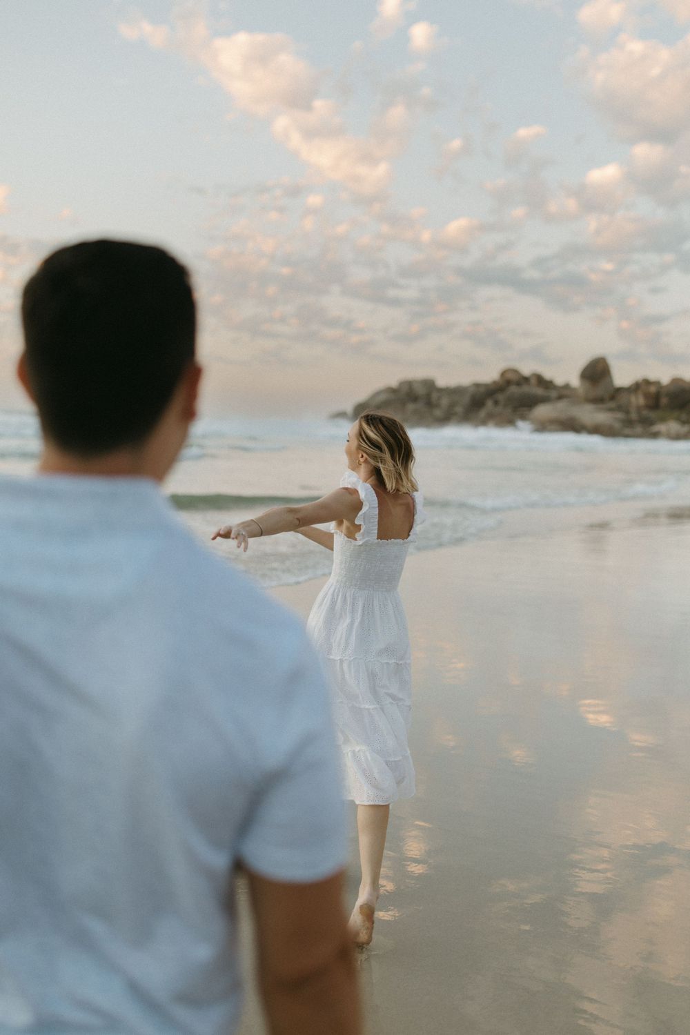 Couple holding hands on a sandy beach at sunset, woman in white dress walking toward ocean waves.