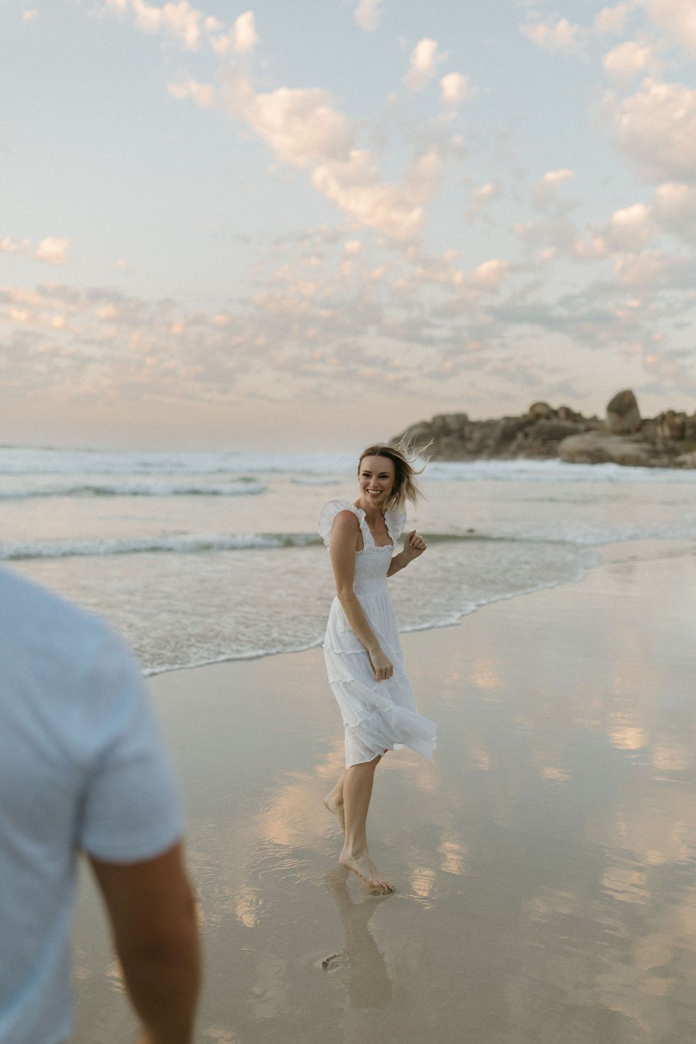 Smiling woman in white dress walking barefoot on wet sand beach at golden hour sunset.