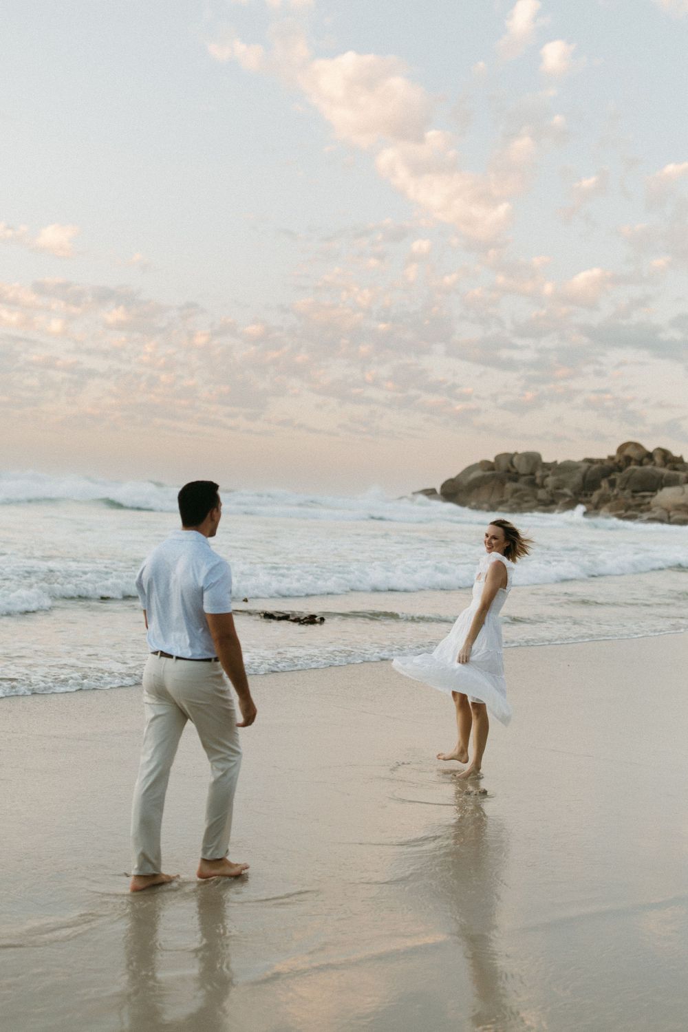 Couple holding hands on a sandy beach at sunset, woman in white dress, waves and rocky shore in background.