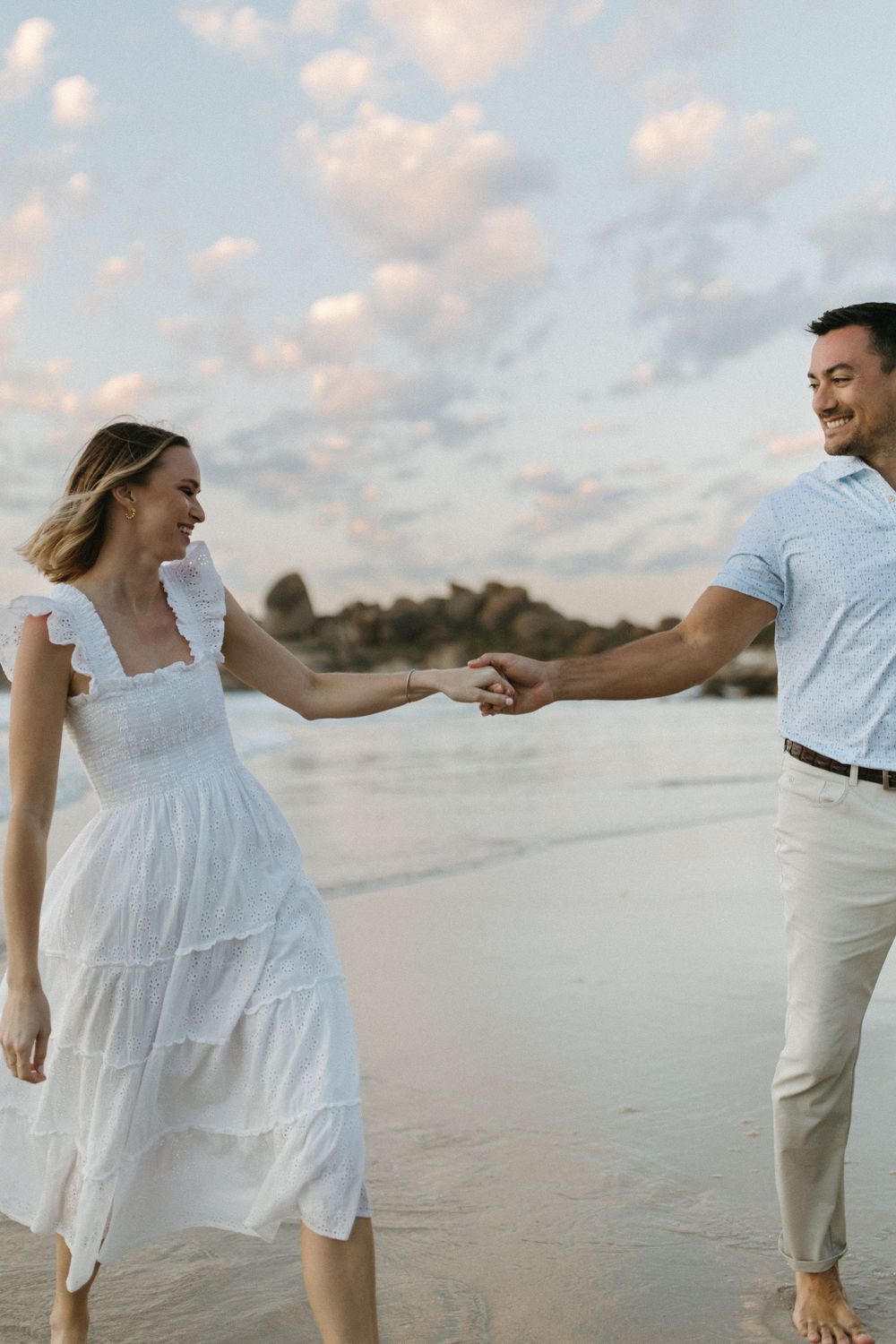 A couple holds hands and smiles while walking barefoot on a beach at sunset, woman in white dress.