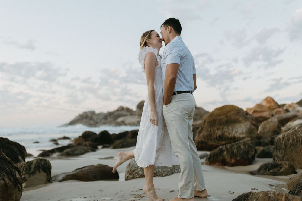 Romantic couple sharing a tender moment on a rocky beach at sunset, woman in white dress, man in light blue shirt.