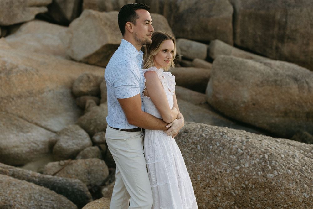 Couple in white outfits embracing near rocky shore, romantic engagement photo session outdoors.
